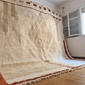 May include: Large, off-white rug with a geometric diamond pattern in shades of brown and tan. The rug has a textured surface and a border with tassels. The rug is displayed indoors, leaning against a wall.