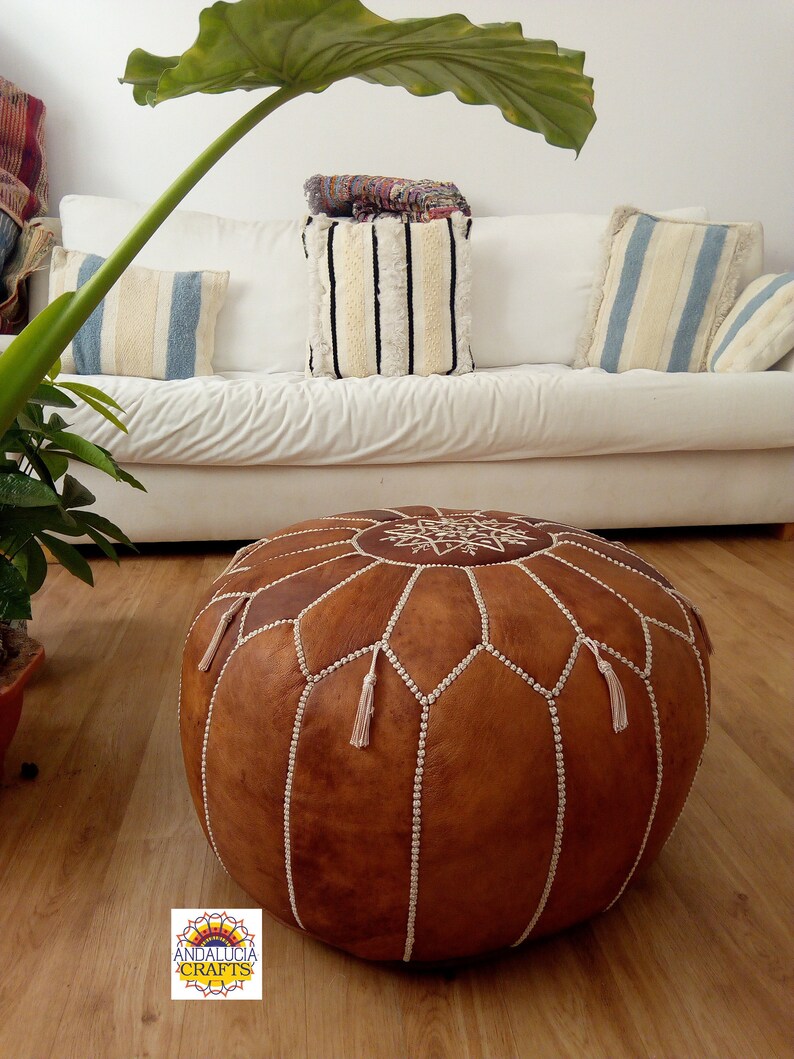 May include: A brown leather pouf with white stitching and tassels. The pouf is sitting on a wooden floor.