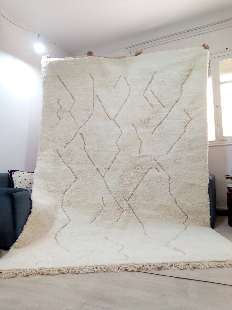 Puede incluir: Una alfombra marroqu&iacute; blanca con un patr&oacute;n geom&eacute;trico en un tono de blanco m&aacute;s claro. La alfombra tiene un borde de flecos.