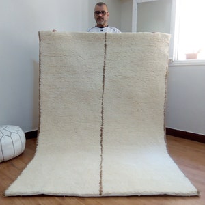 Puede incluir: Una alfombra blanca de pelo largo con una franja marr&oacute;n en el centro. La alfombra est&aacute; doblada por la mitad y una persona la sostiene.