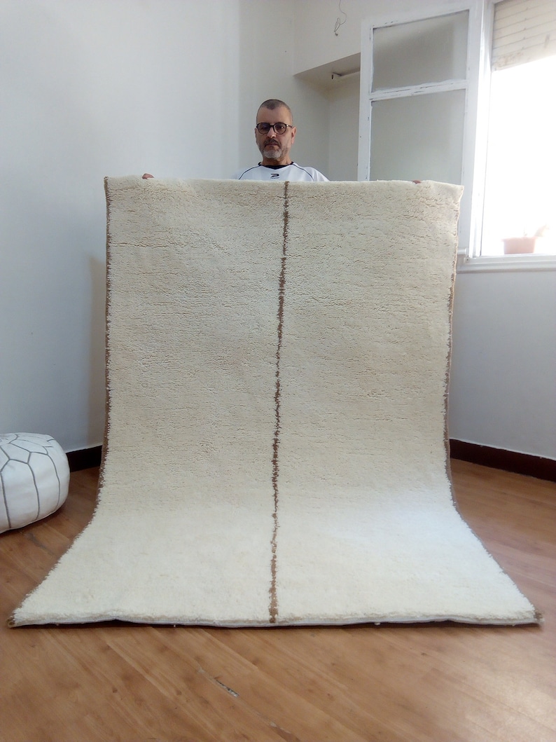 Puede incluir: Una alfombra blanca de pelo largo con una franja marr&oacute;n en el centro. La alfombra est&aacute; doblada por la mitad y una persona la sostiene.