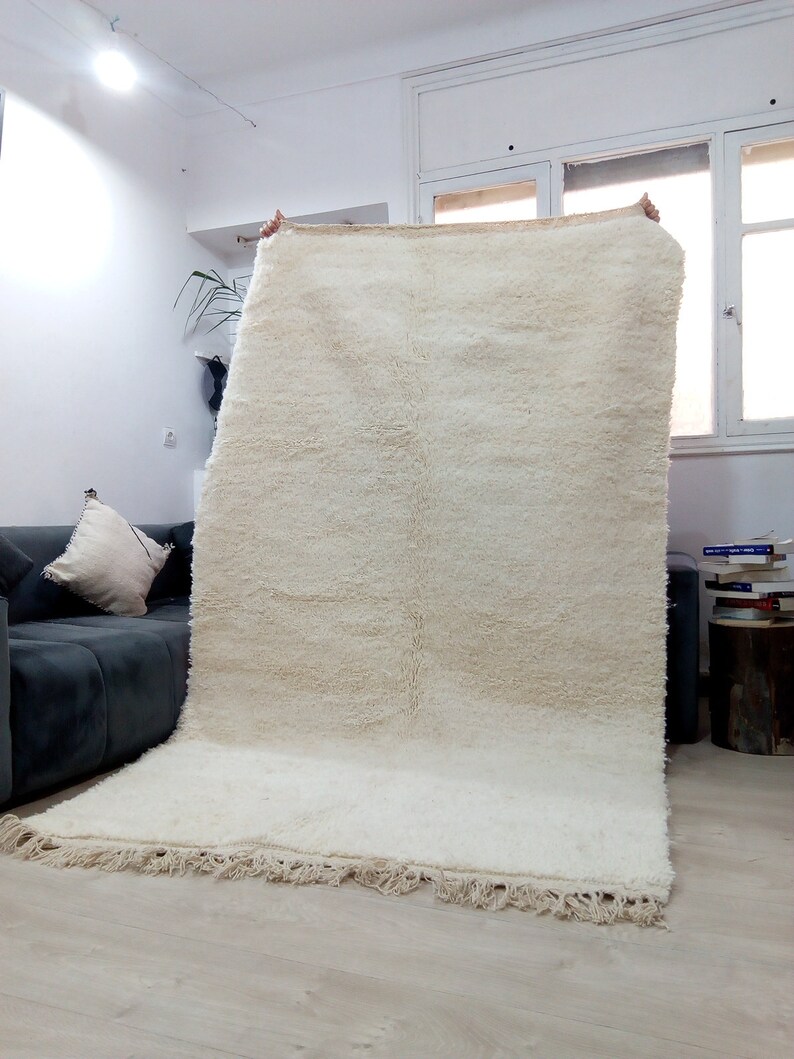 Pode incluir: Um tapete branco e felpudo, tecido &agrave; m&atilde;o, com franjas nas bordas. O tapete est&aacute; sendo segurado por duas m&atilde;os.