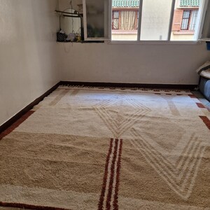 May include: A large, rectangular rug with a geometric design. The rug features a cream-colored background with brown and red accents. The design includes stripes and chevron patterns, with a fringed edge.