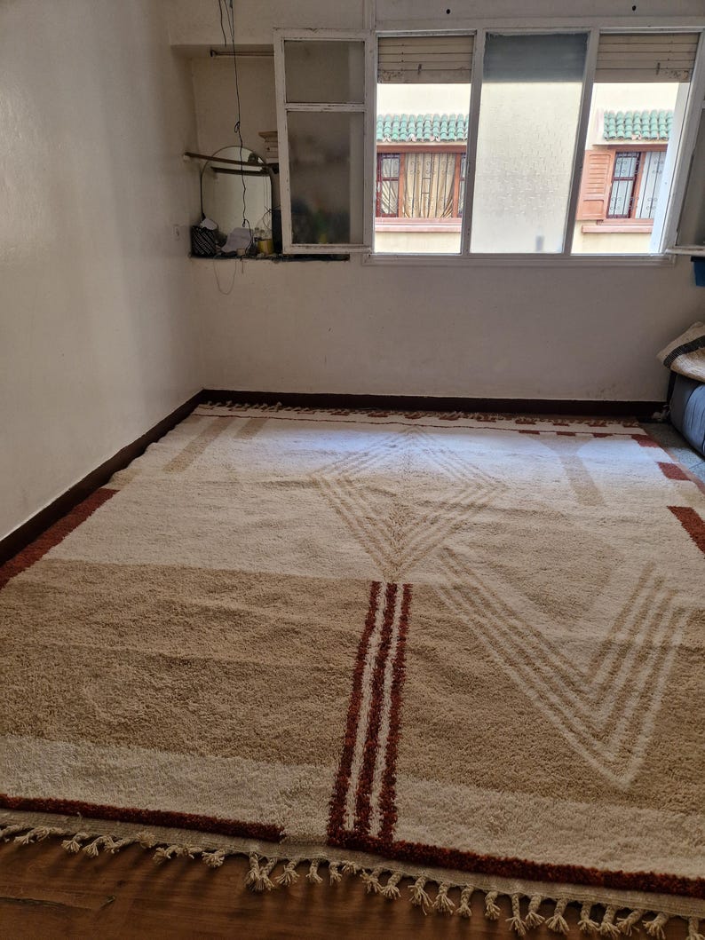 May include: A large, rectangular rug with a geometric design. The rug features a cream-colored background with brown and red accents. The design includes stripes and chevron patterns, with a fringed edge.