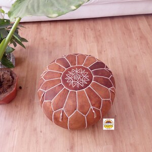 May include: A brown leather pouf with white stitching and a star design. The pouf is sitting on a wooden floor.