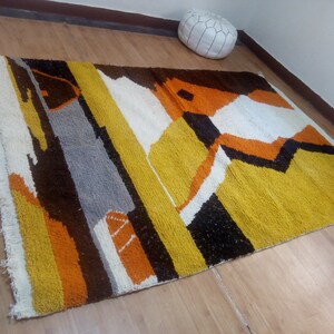 May include: A rectangular rug with a geometric design. The rug features a combination of colors, including yellow, brown, orange, white, gray, and black. The rug has a fringed edge on one side.