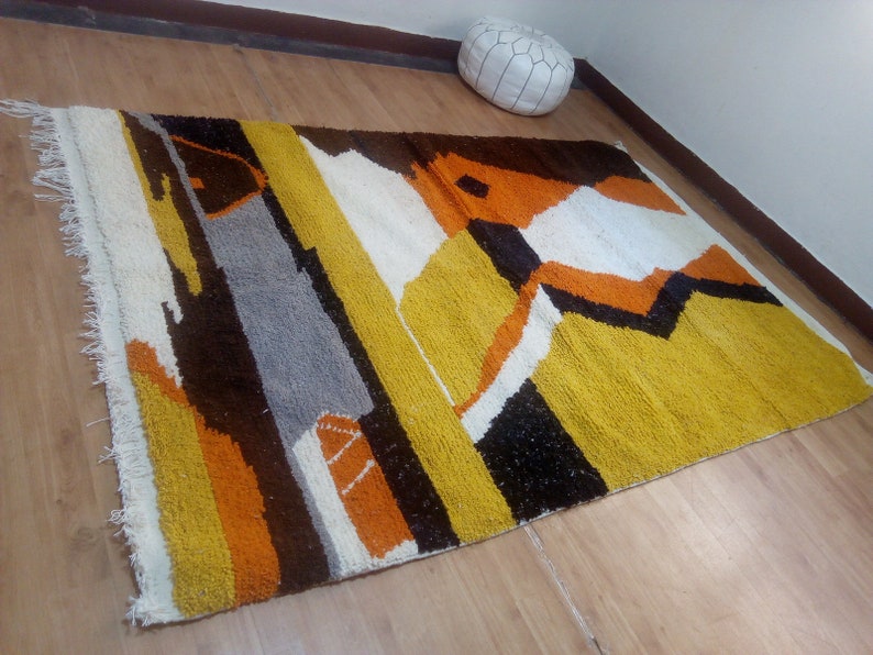 May include: A rectangular rug with a geometric design. The rug features a combination of colors, including yellow, brown, orange, white, gray, and black. The rug has a fringed edge on one side.