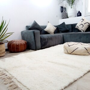 Pode incluir: Um tapete branco de pelo alto com franjas nas bordas, colocado em um piso de madeira em uma sala de estar. O tapete est&aacute; na frente de um sof&aacute; de canto cinza com almofadas e um pufe de couro marrom.