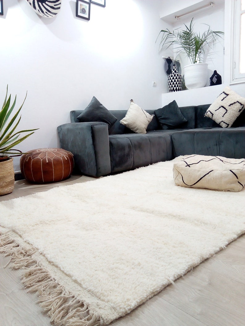 Pode incluir: Um tapete branco de pelo alto com franjas nas bordas, colocado em um piso de madeira em uma sala de estar. O tapete est&aacute; na frente de um sof&aacute; de canto cinza com almofadas e um pufe de couro marrom.
