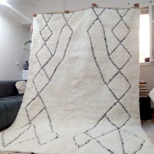 May include: A white Moroccan rug with a black diamond pattern. The rug has a fringe border and is being held up by two people.