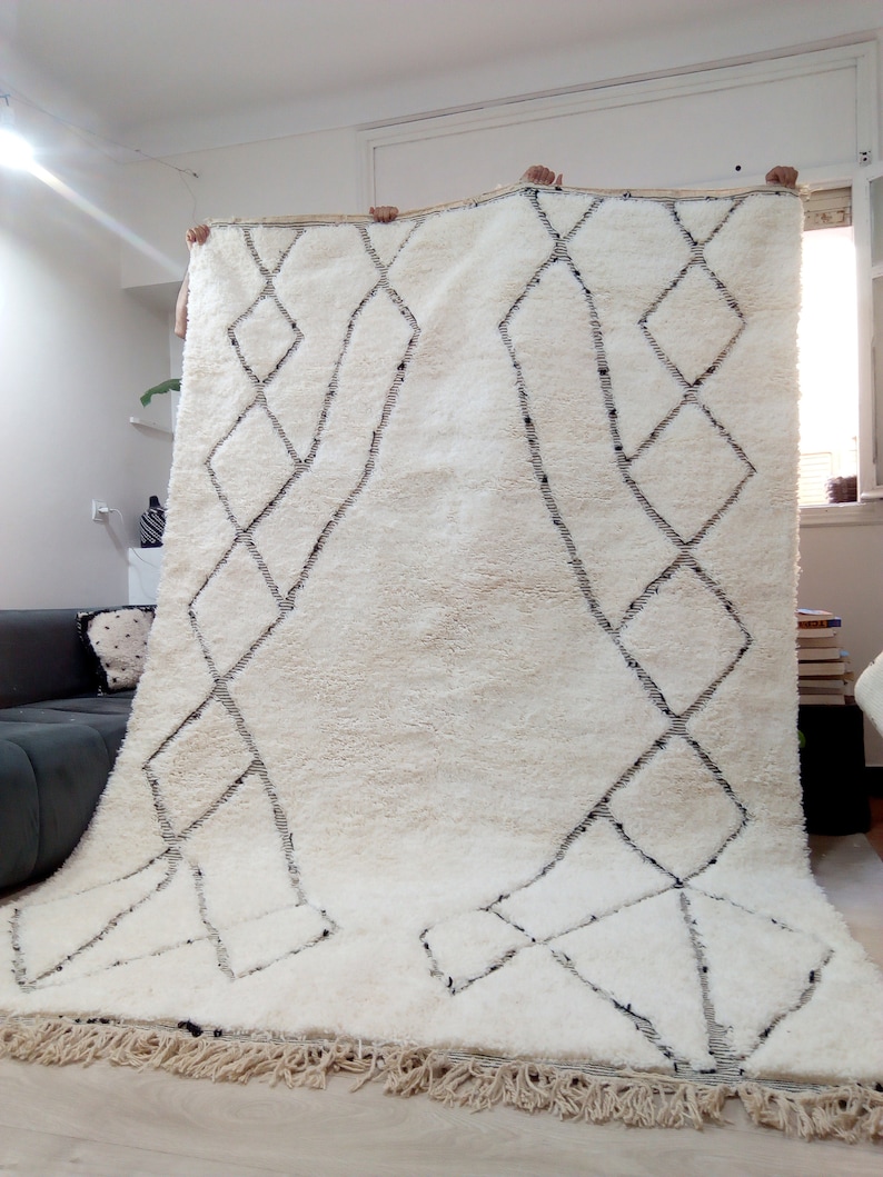 May include: A white Moroccan rug with a black diamond pattern. The rug has a fringe border and is being held up by two people.