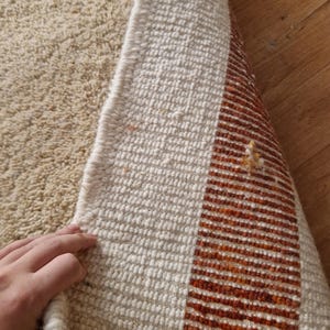 May include: Beige and off-white woven rug with a geometric design. The rug features a textured surface and a border with orange and brown stripes. The rug is partially rolled up, revealing the underside.