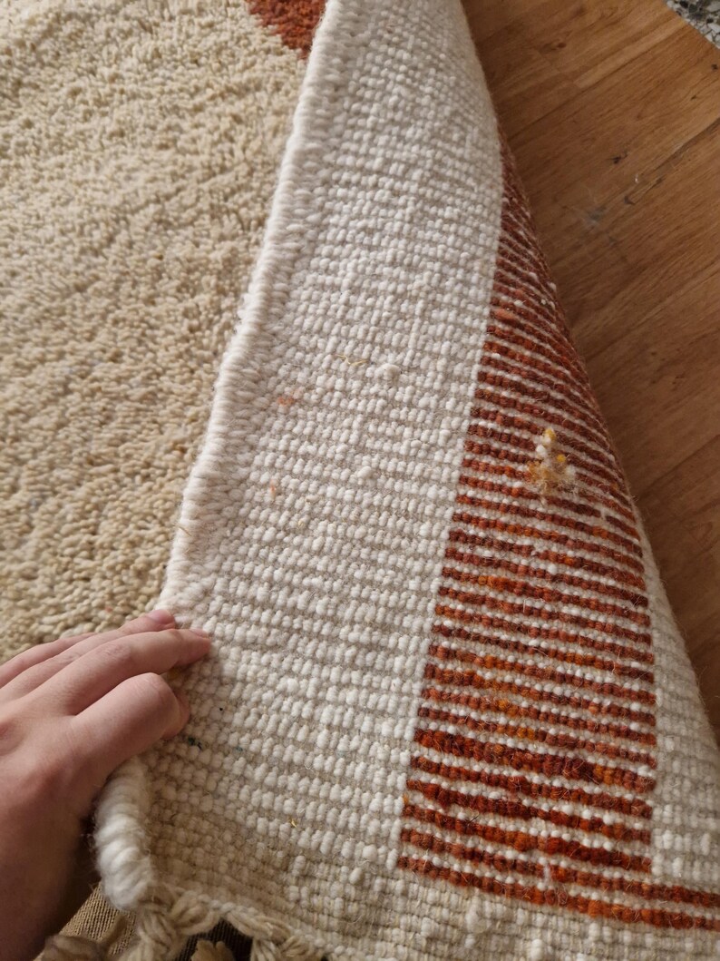 May include: Beige and off-white woven rug with a geometric design. The rug features a textured surface and a border with orange and brown stripes. The rug is partially rolled up, revealing the underside.