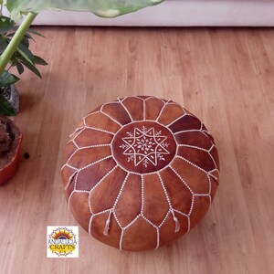May include: A brown leather pouf with white stitching and a star design. The pouf is sitting on a wooden floor.