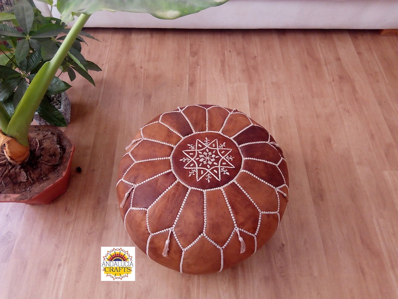 May include: A brown leather pouf with white stitching and a star design. The pouf is sitting on a wooden floor.