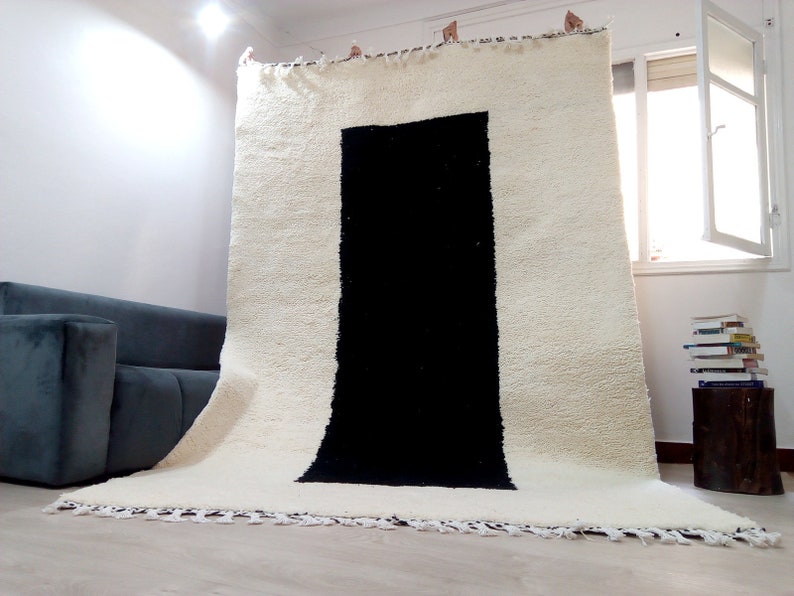 May include: A white and black Moroccan rug with a large black rectangle in the center. The rug has a fringe border and is hanging on a wall.