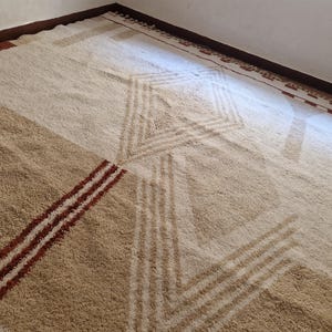 May include: A large, geometric patterned rug in shades of beige, tan, and brown. The rug features a central diamond design and diagonal stripes, creating a modern, textured look. The rug is placed in a corner of a room.
