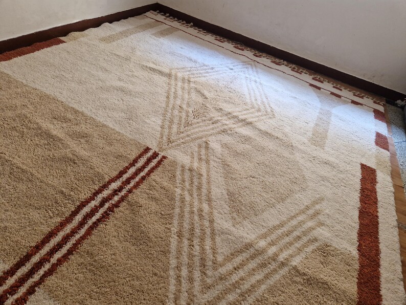 May include: A large, geometric patterned rug in shades of beige, tan, and brown. The rug features a central diamond design and diagonal stripes, creating a modern, textured look. The rug is placed in a corner of a room.