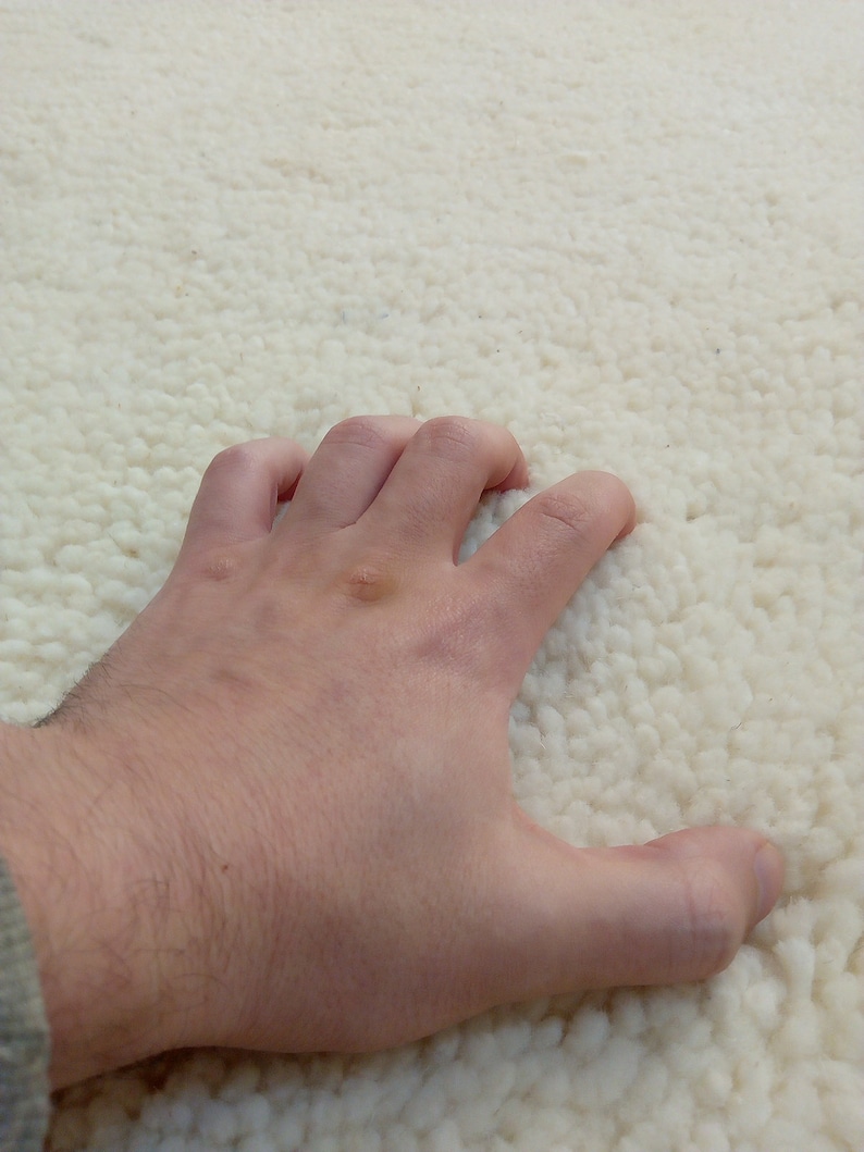 Puede incluir: Primer plano de una mano tocando una alfombra blanca y esponjosa. La alfombra tiene una textura suave y mullida.