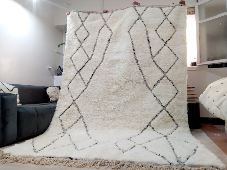 May include: A white Moroccan rug with a black diamond pattern. The rug has a fringe border and is made of wool.