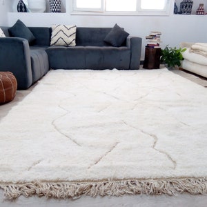 Puede incluir: Una alfombra blanca de pelo largo con un patr&oacute;n geom&eacute;trico y bordes con flecos. La alfombra est&aacute; en un sal&oacute;n con un sof&aacute; de esquina gris y un sof&aacute; blanco.