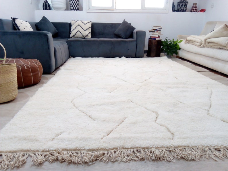 Puede incluir: Una alfombra blanca de pelo largo con un patr&oacute;n geom&eacute;trico y bordes con flecos. La alfombra est&aacute; en un sal&oacute;n con un sof&aacute; de esquina gris y un sof&aacute; blanco.