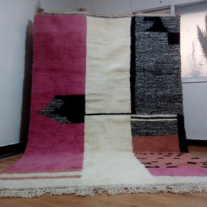 May include: A hand-woven rug with a geometric design in shades of pink, white, and black. The rug has a fringe border.