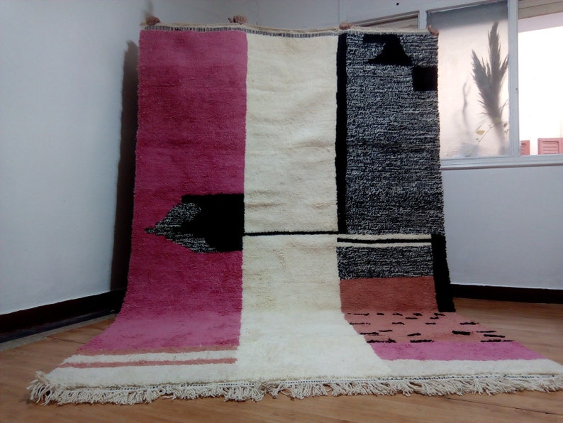 May include: A hand-woven rug with a geometric design in shades of pink, white, and black. The rug has a fringe border.