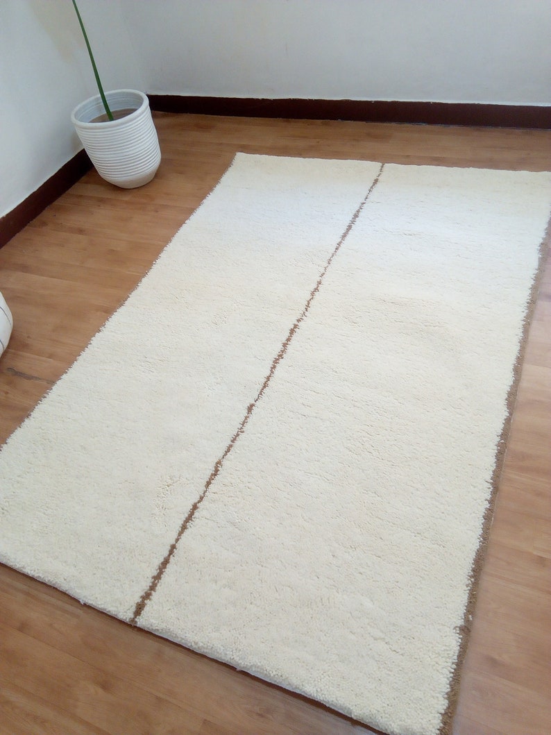 Puede incluir: Una alfombra de pelo blanco con una franja marr&oacute;n en el centro. La alfombra est&aacute; sobre un suelo de madera.
