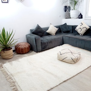 Pode incluir: Um tapete felpudo branco com franjas nas bordas, um pufe de couro marrom e um pufe com padr&atilde;o branco e preto em um piso de madeira clara. Um sof&aacute; de canto cinza com almofadas brancas est&aacute; no fundo.