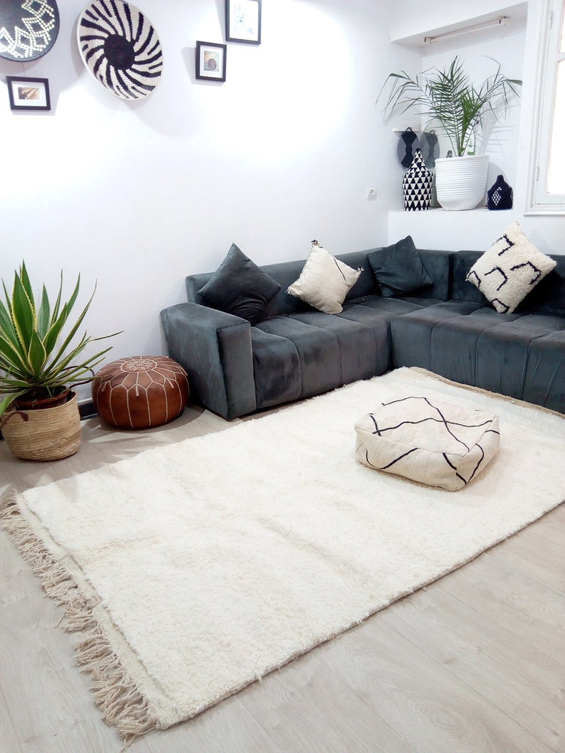 Pode incluir: Um tapete felpudo branco com franjas nas bordas, um pufe de couro marrom e um pufe com padr&atilde;o branco e preto em um piso de madeira clara. Um sof&aacute; de canto cinza com almofadas brancas est&aacute; no fundo.