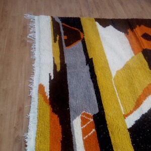 May include: A colorful, geometric rug with a textured surface. The rug features a design of vertical stripes and shapes in shades of yellow, orange, gray, white, and black. The rug has a fringed edge.