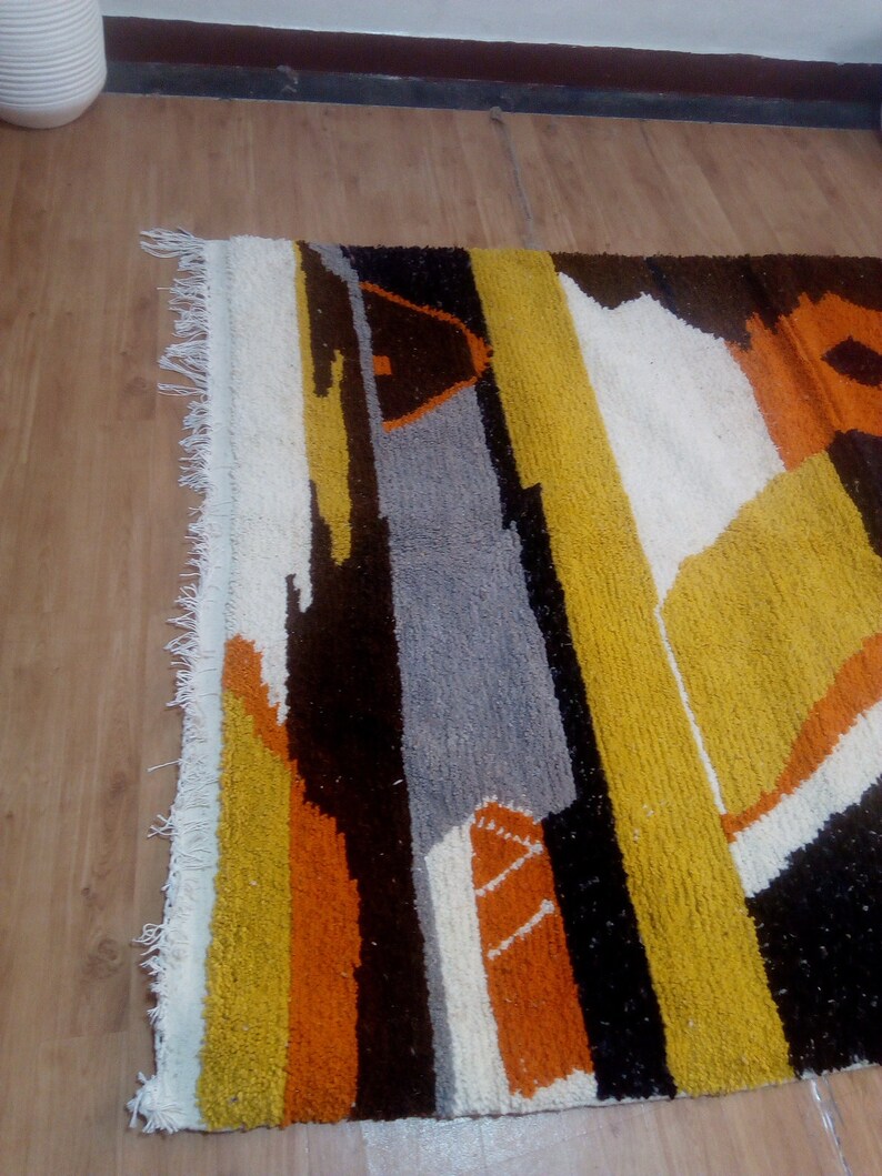 May include: A colorful, geometric rug with a textured surface. The rug features a design of vertical stripes and shapes in shades of yellow, orange, gray, white, and black. The rug has a fringed edge.