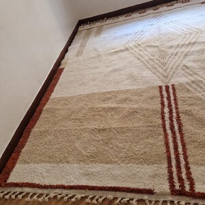 May include: A rectangular, beige and brown area rug with geometric patterns. The rug features a central diamond design, horizontal stripes, and vertical red stripes. The rug has a fringe along the bottom edge. The rug is made of a soft, textured material.