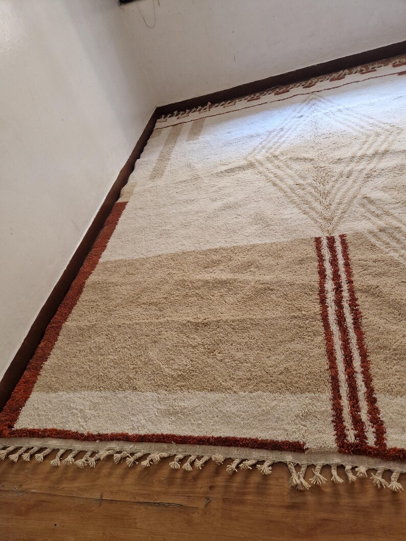 May include: A rectangular, beige and brown area rug with geometric patterns. The rug features a central diamond design, horizontal stripes, and vertical red stripes. The rug has a fringe along the bottom edge. The rug is made of a soft, textured material.