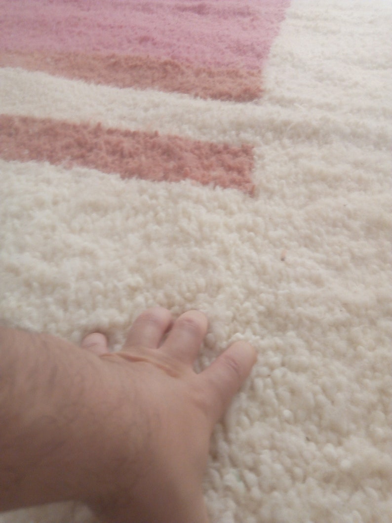 May include: A close-up of a white fluffy rug with pink and peach stripes. A hand is resting on the rug.