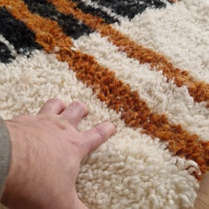 May include: Close-up of a white, orange, and black striped rug with a hand resting on the white portion. The rug has a thick, fluffy texture.