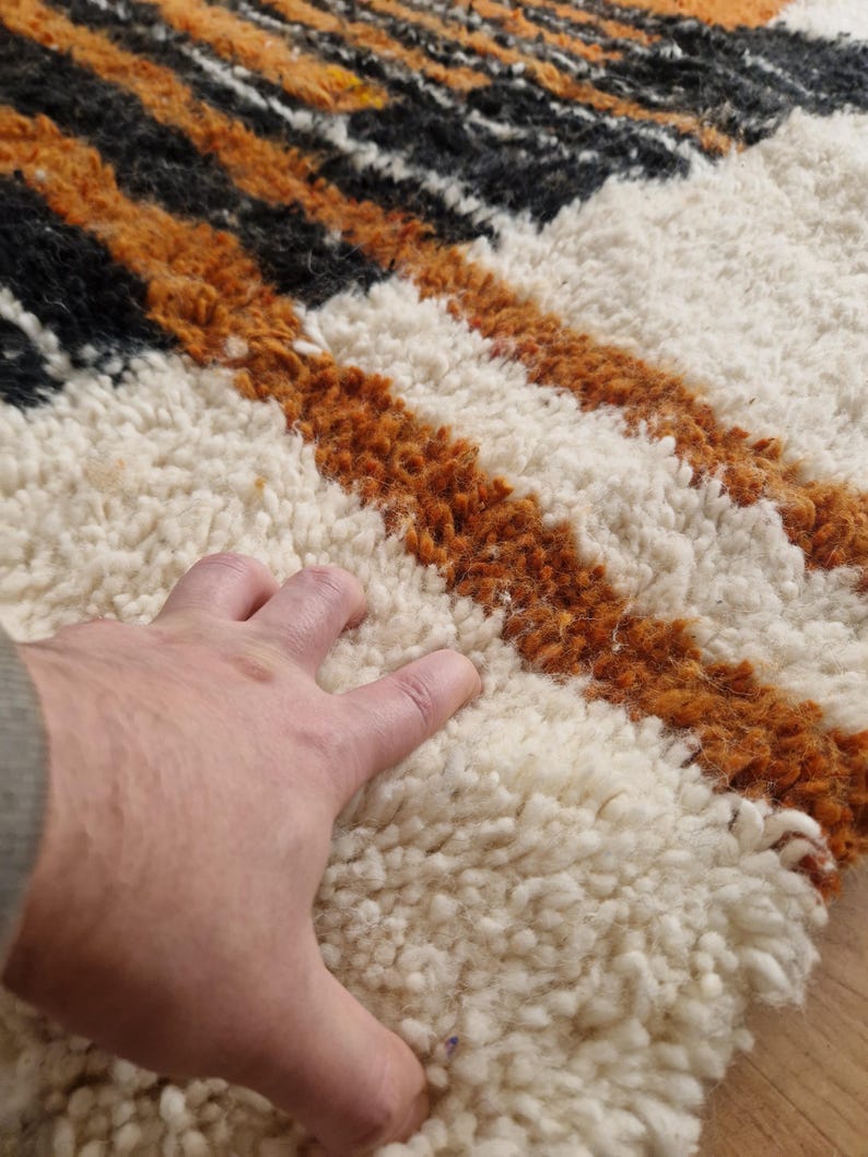 May include: Close-up of a white, orange, and black striped rug with a hand resting on the white portion. The rug has a thick, fluffy texture.
