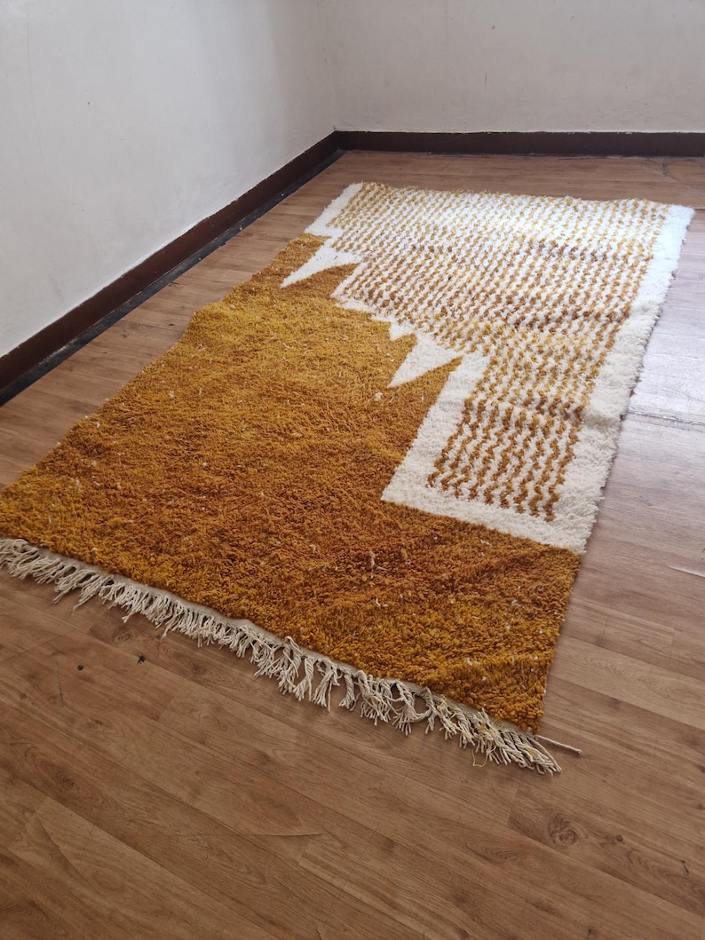 May include: A mustard yellow and white geometric patterned rug with fringe. The rug has a modern design with a white geometric pattern on a mustard yellow background.