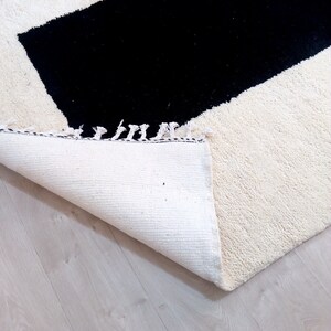 May include: A white rug with a black rectangular shape in the center. The rug has a fringe border and is folded over on a light wood floor.