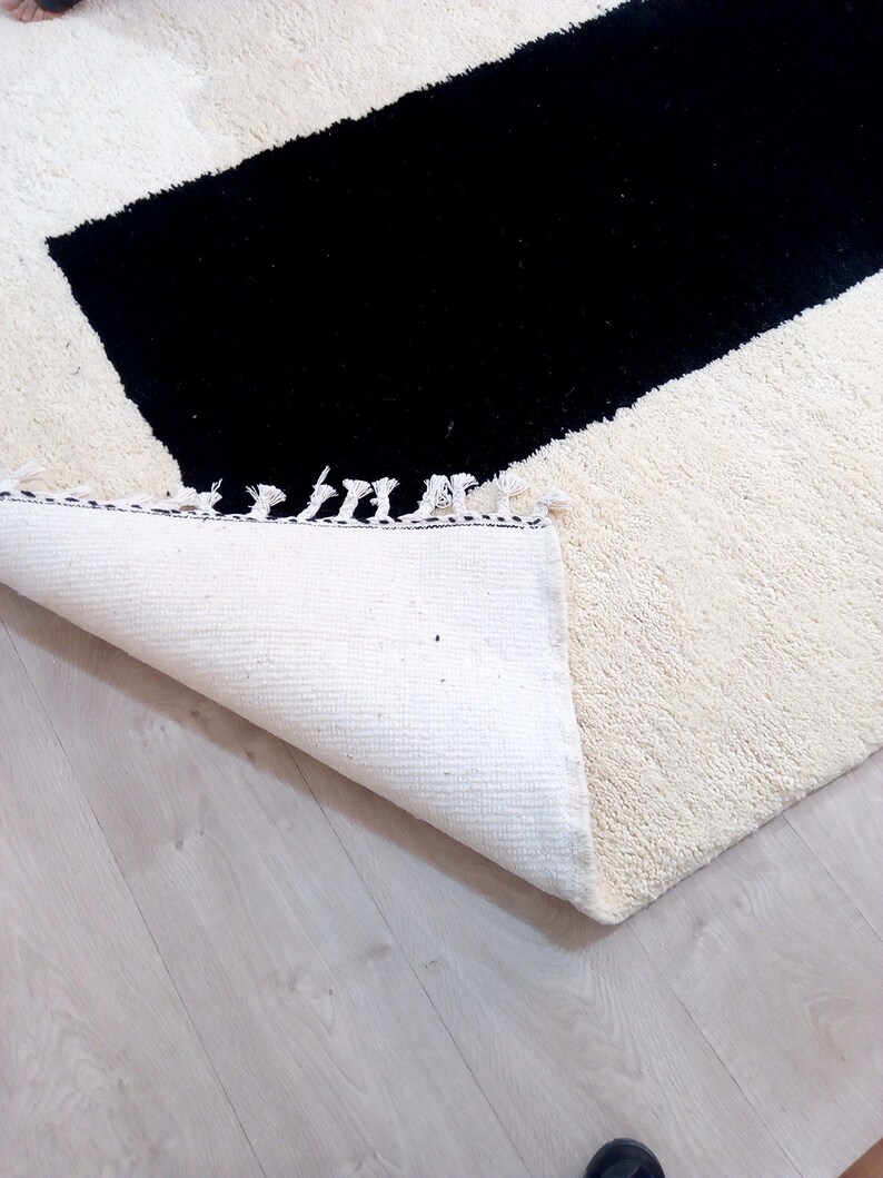 May include: A white rug with a black rectangular shape in the center. The rug has a fringe border and is folded over on a light wood floor.