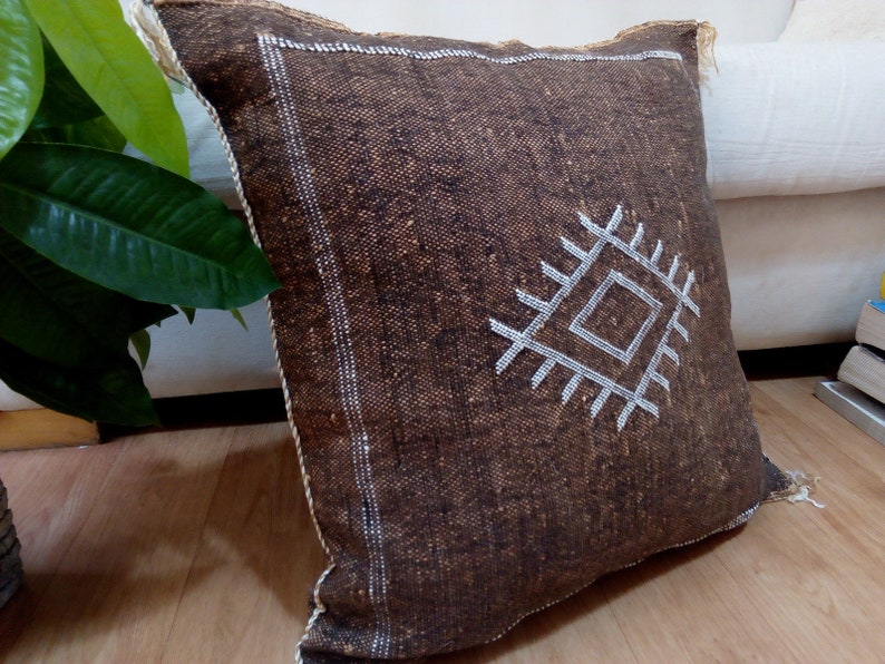 Puede incluir: Una almohada cuadrada marr&oacute;n con un dise&ntilde;o geom&eacute;trico blanco. La almohada est&aacute; hecha de una tela texturizada y tiene un borde de flecos.