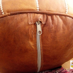 May include: A brown leather pouf with a zipper closure. The pouf is made of stitched leather panels and has a white cord detail.