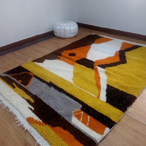 May include: A rectangular rug with a geometric design in shades of yellow, orange, brown, gray, and white. The rug has a textured surface and a fringed edge. The rug is placed on a wooden floor.