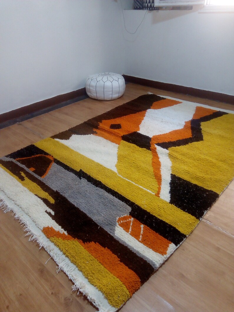 May include: A rectangular rug with a geometric design in shades of yellow, orange, brown, gray, and white. The rug has a textured surface and a fringed edge. The rug is placed on a wooden floor.