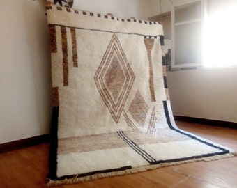 May include: A white, brown, and black Moroccan rug with a geometric pattern. The rug has a fringe border and is made of wool.
