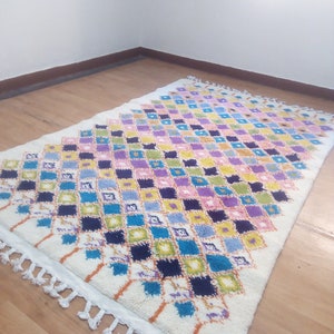 May include: A white Moroccan rug with a diamond pattern in shades of blue, green, yellow, pink, and purple. The rug has a white fringe border.