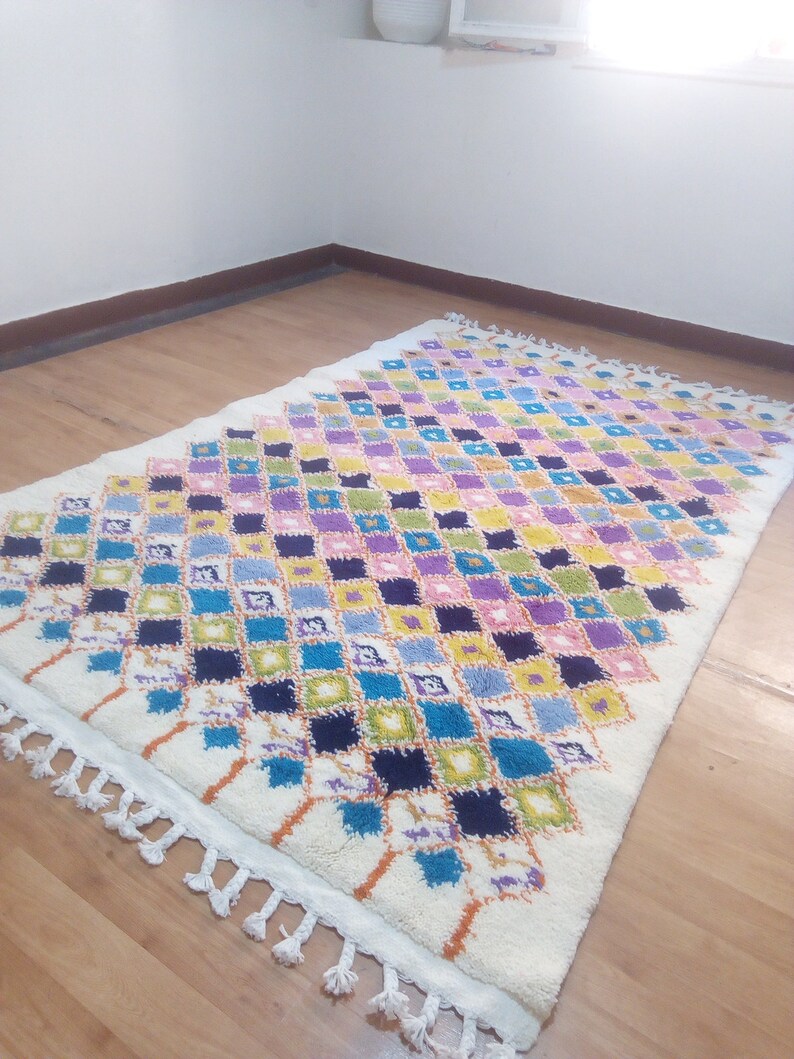 May include: A white Moroccan rug with a diamond pattern in shades of blue, green, yellow, pink, and purple. The rug has a white fringe border.