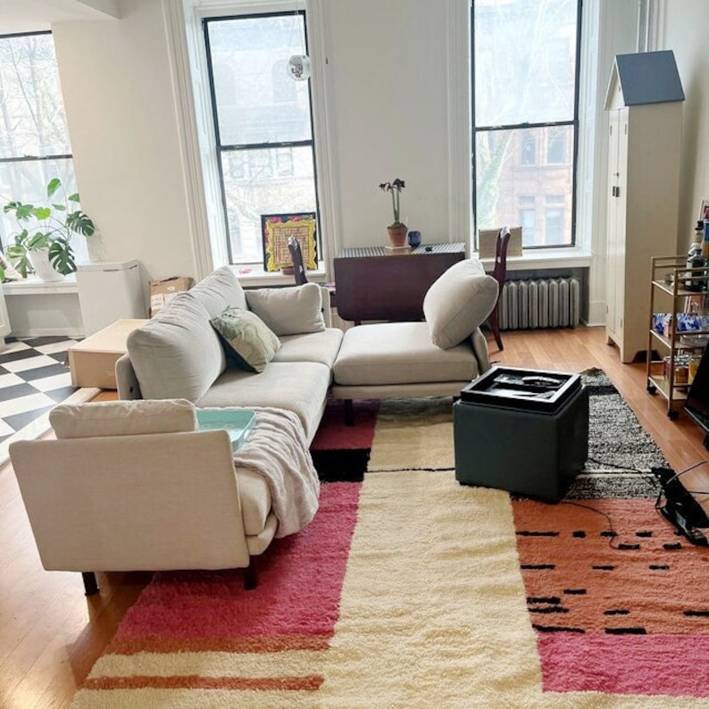 May include: A light gray sectional sofa with a light gray throw blanket on the armrest. The sofa is in a living room with a rug in shades of pink, beige, and black. There is a small black storage ottoman in front of the sofa.