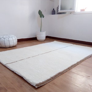 Puede incluir: Una alfombra de pelo blanco con una franja marr&oacute;n en el centro. La alfombra est&aacute; sobre un suelo de madera.
